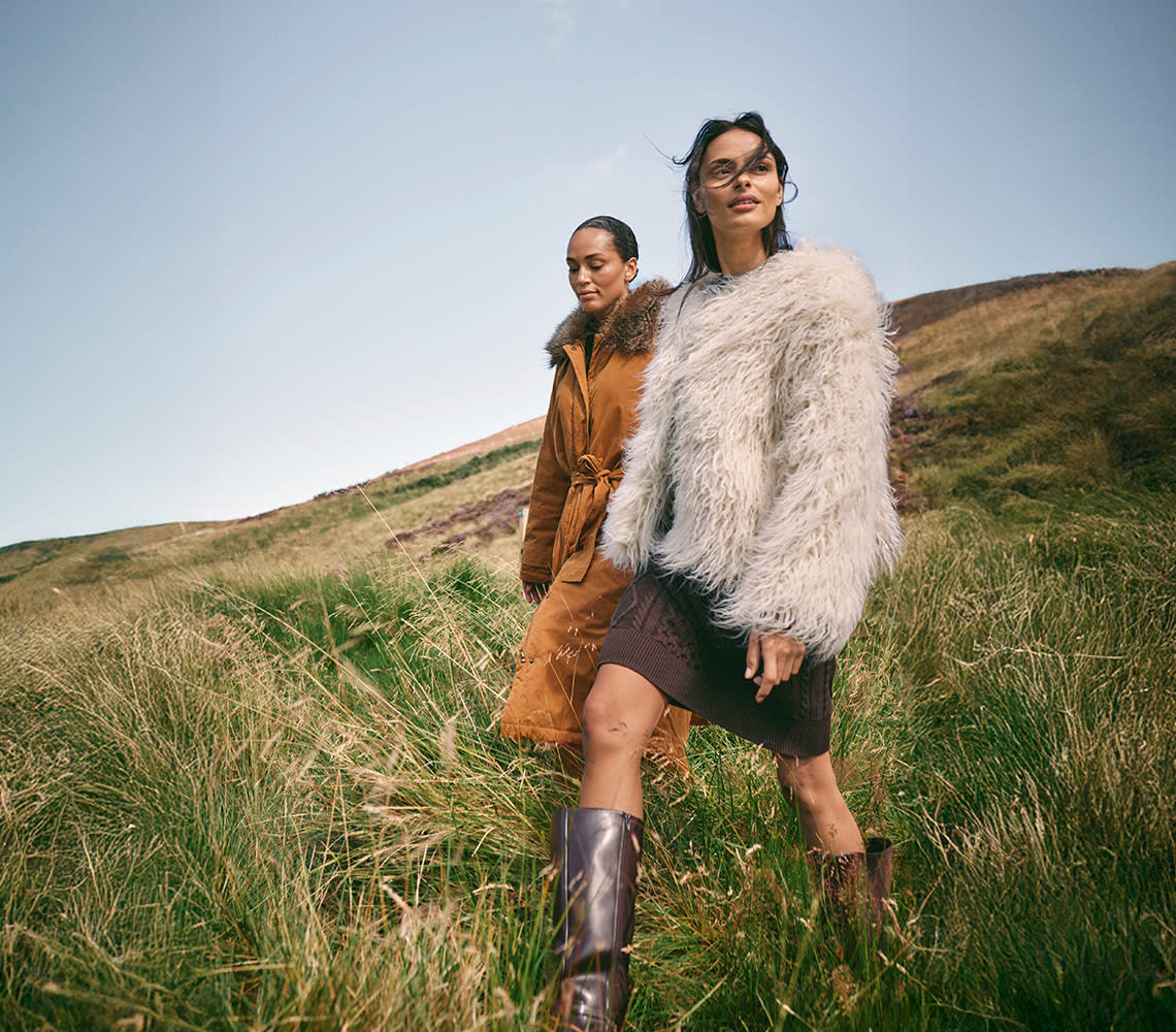 Two women in fur coats walk side by side through a lush green field, showcasing bonprix clothing.